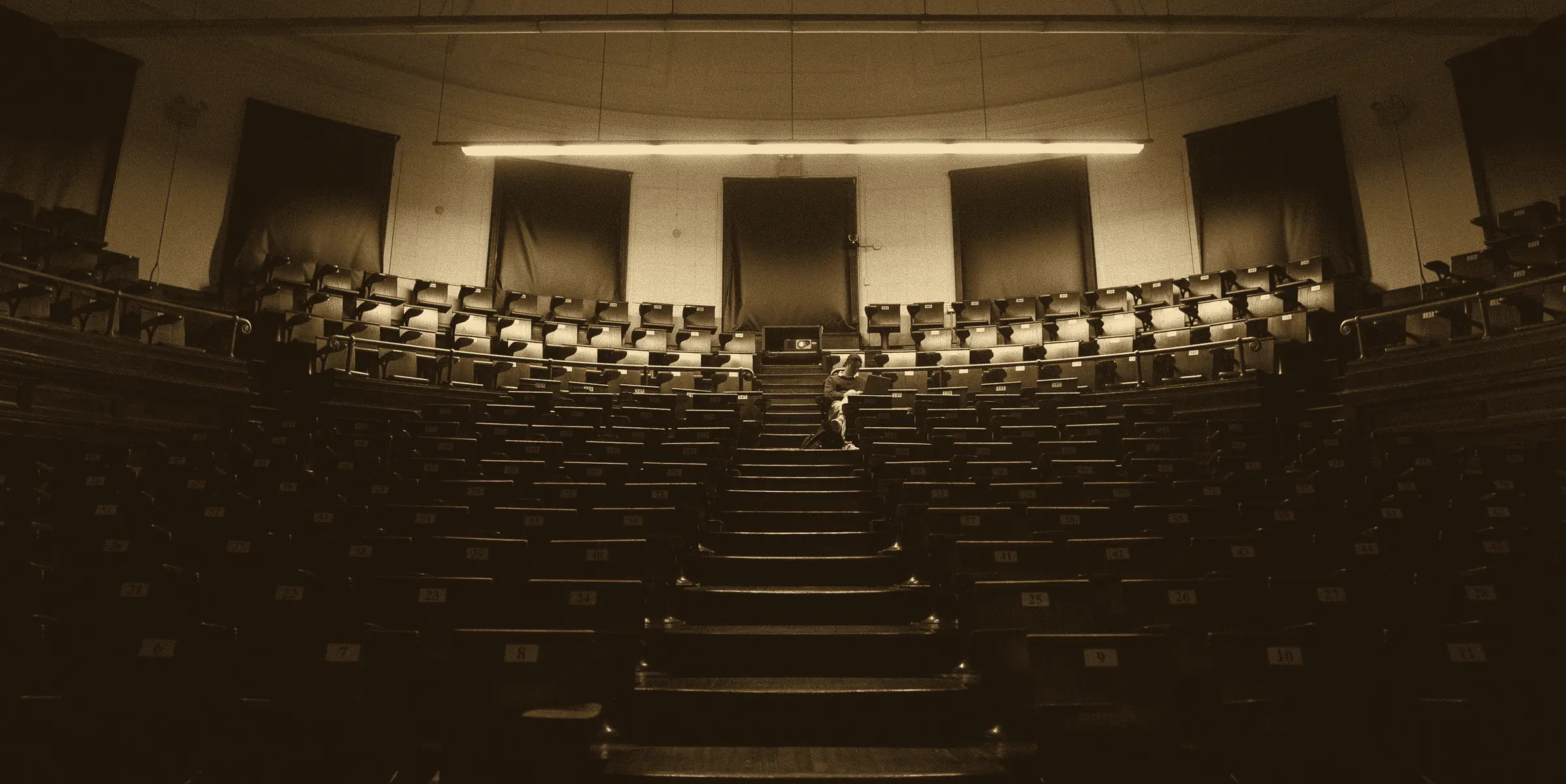 An empty lecture theatre: tiers of wooden seats rise toward a single strip light, and one student sits reading on the stairs.
